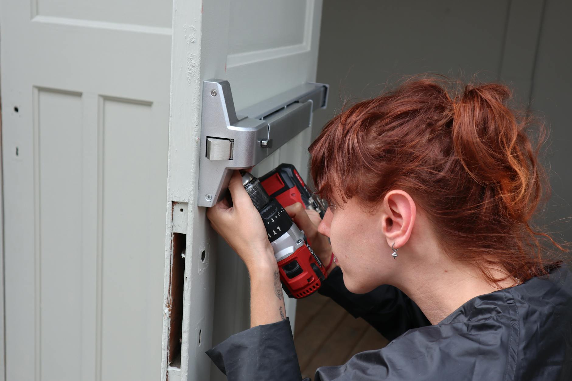 A woman with red hair uses a drill to fix a door handle.