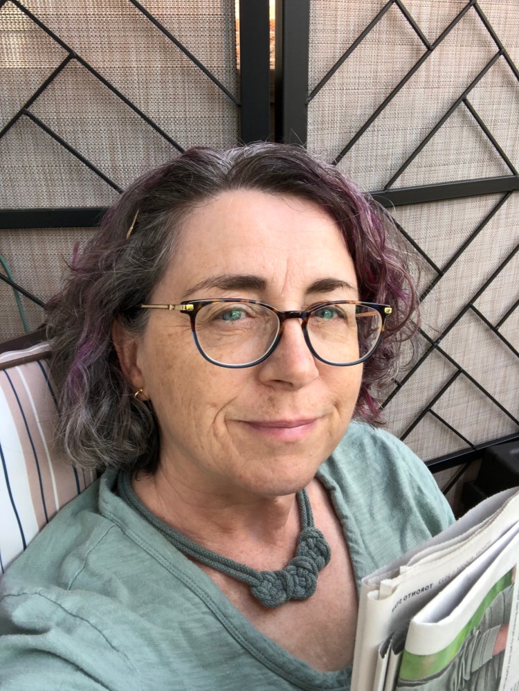 The author wearing a green t-shirt and green necklace, sitting outside reading the newspaper and lacking enthusiasm.