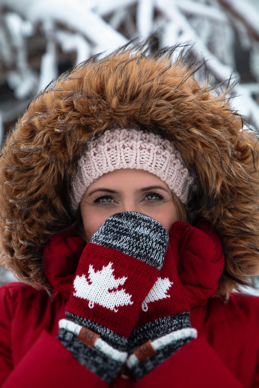 A woman wearing a red winter coat with the fur hood pulled over her knitted hat is covering her mouth with her red maple leaf mittens.