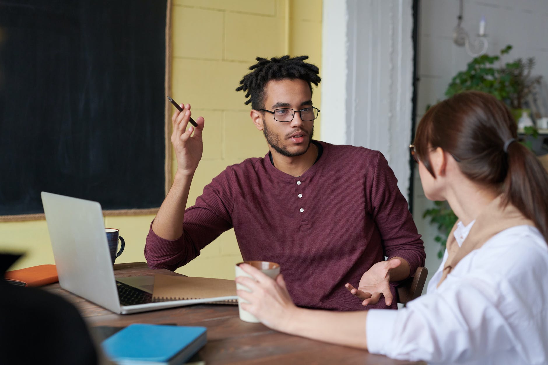 A man using his hands to explain something to another person as they sit casually at a table, with coffee cups and a computer.