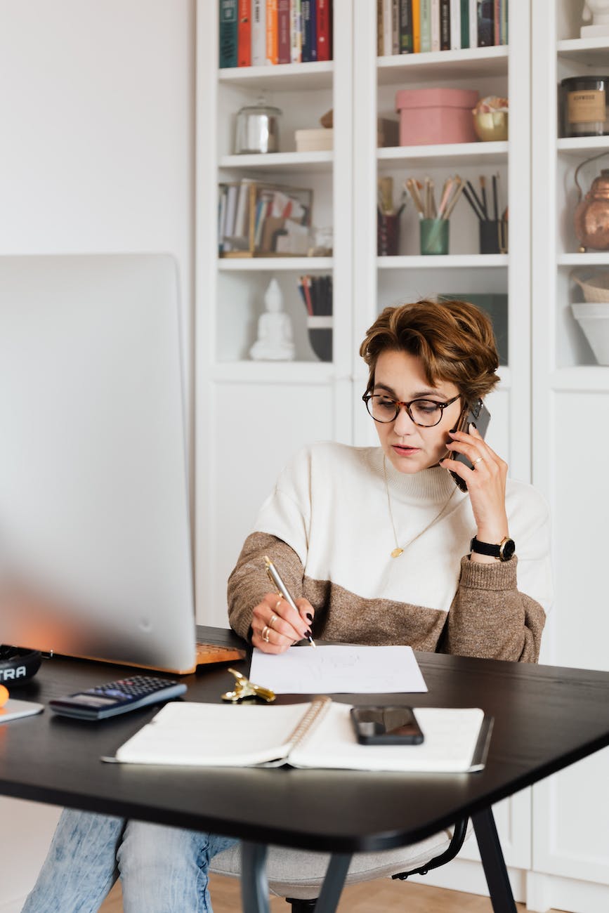 A woman on sitting in front of a computer, on the phone and taking notes on paper.