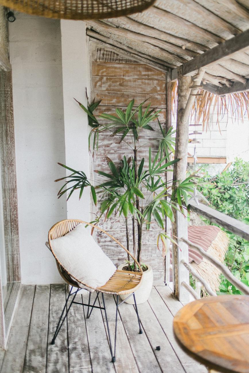 A bamboo chair with cushion is on a wooden balcony. Tropical plants can be seen in a pot and on the ground below.