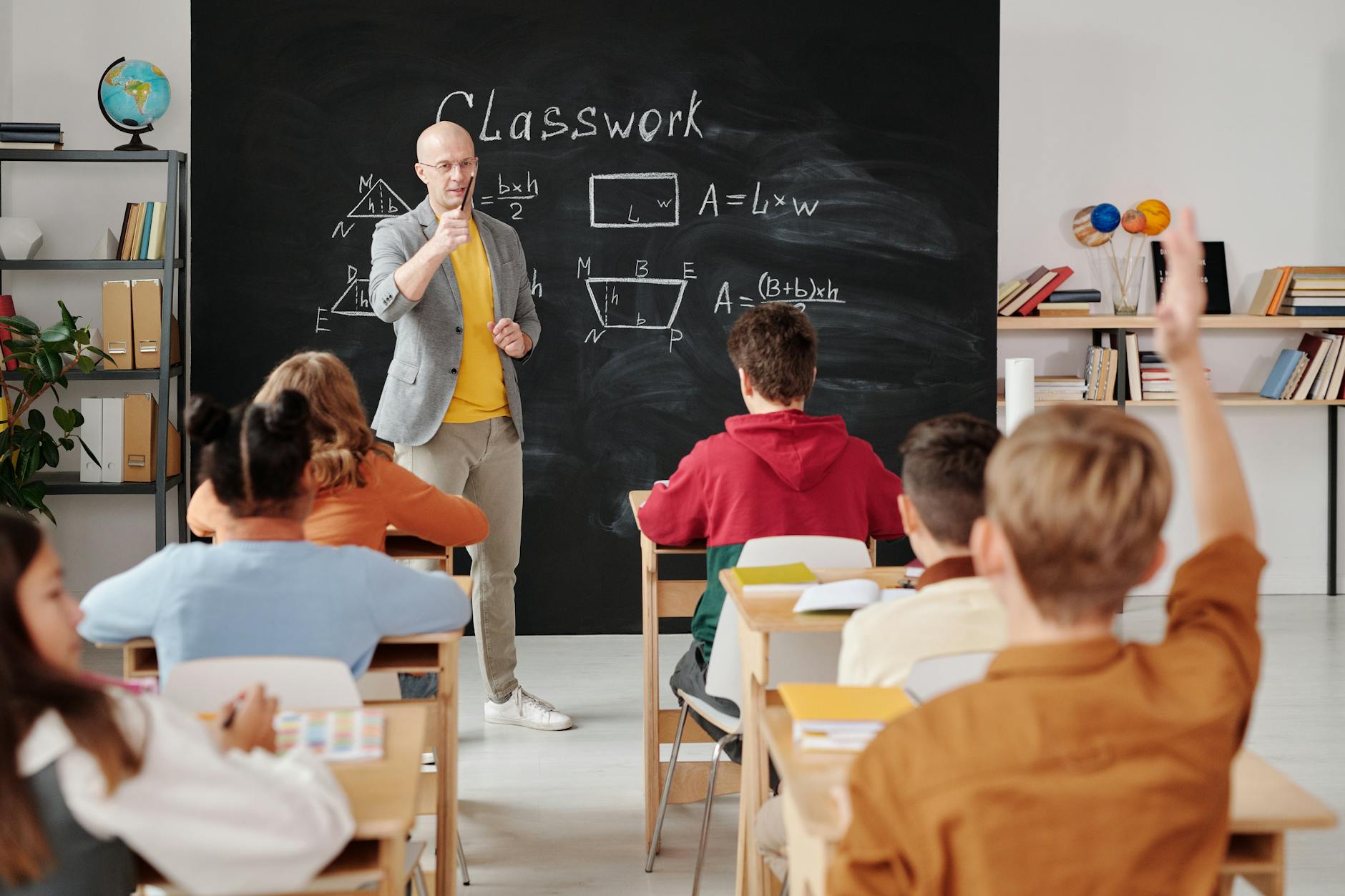 A male teacher stands in front of a chalkboard with math on it. He is calling on a student who has their hand raised.