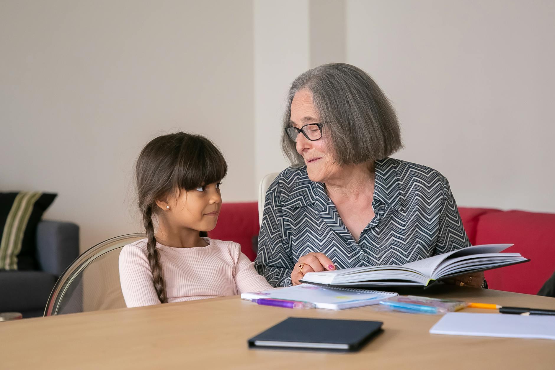 A grandmother is helping a girl with her homework. There are books spread on the table in front of them. 