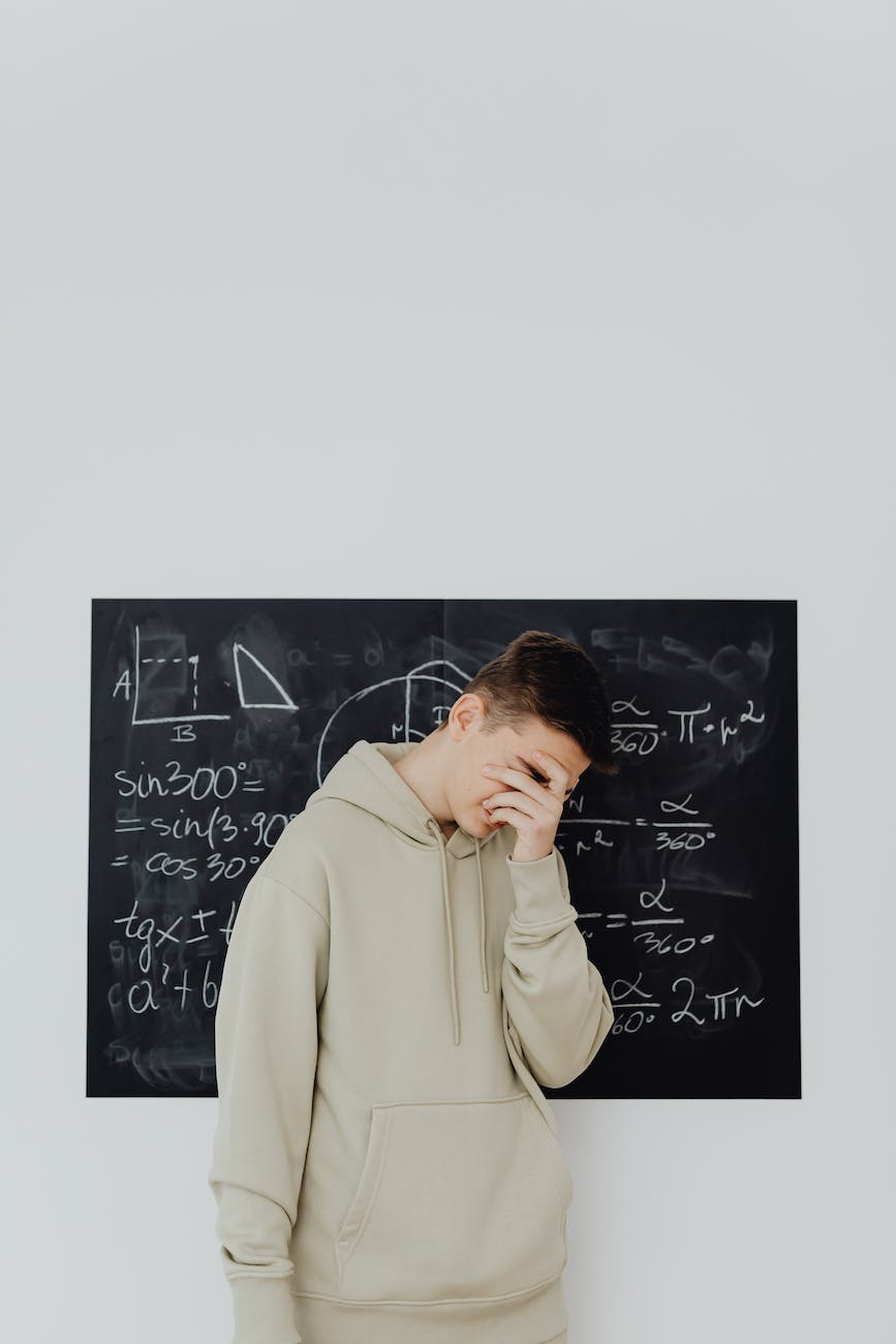 A youth is standing in front of a chalkboard with math on it. He has his hand covering his face in shame.