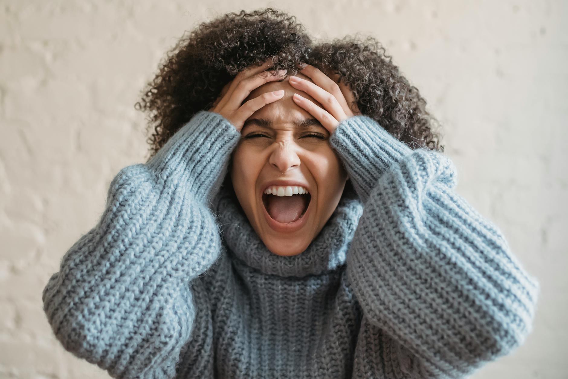 A woman wearing a blue sweater is holding her hands at her temples and yelling in frustration.