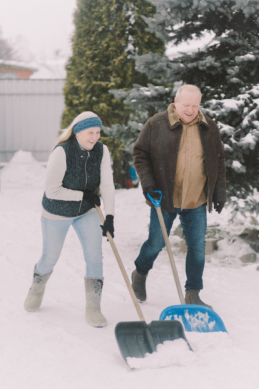 A man and woman are each using a snow shovel as they walk beside each other smiling and talking.