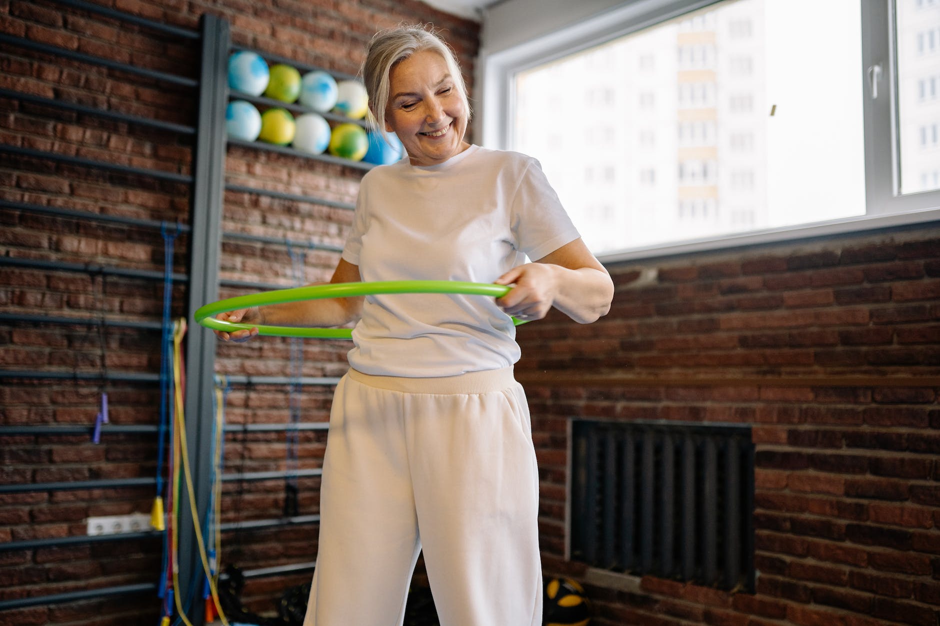 A grey-haired woman smiling while using a hoola-hoop in a gym. She's wearing a white t-shirt and light coloured sweatpants.