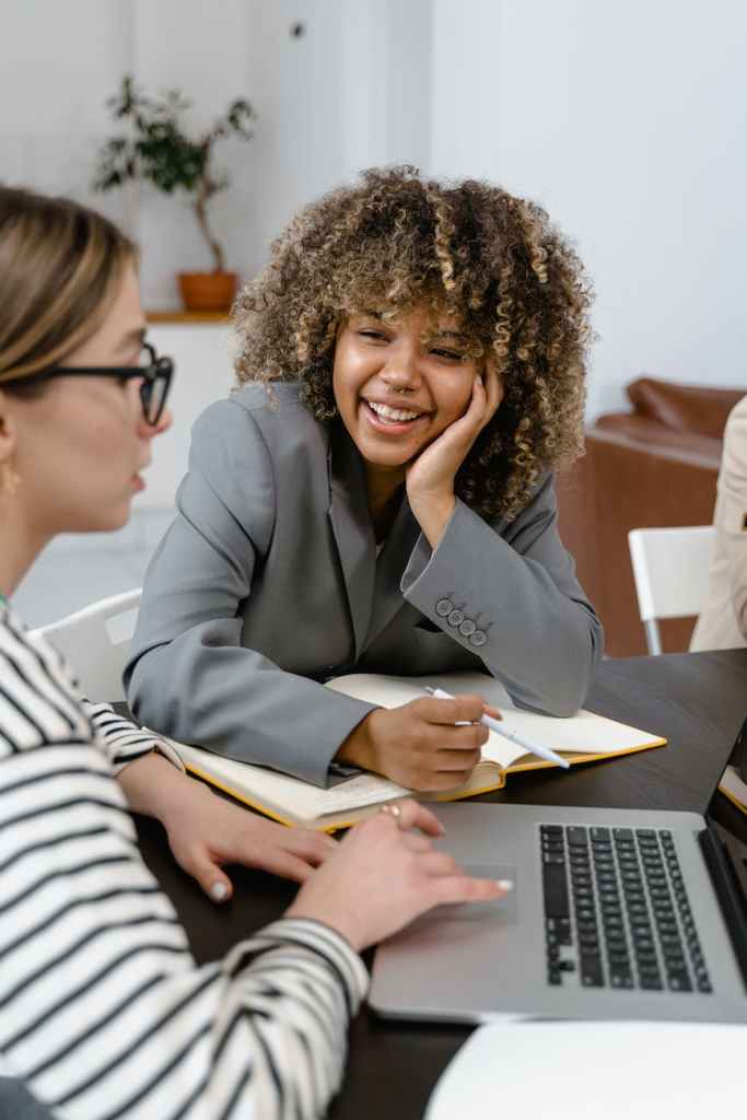 A woman smiling with a book in front of her on the table and another woman is listening while using her computer. 