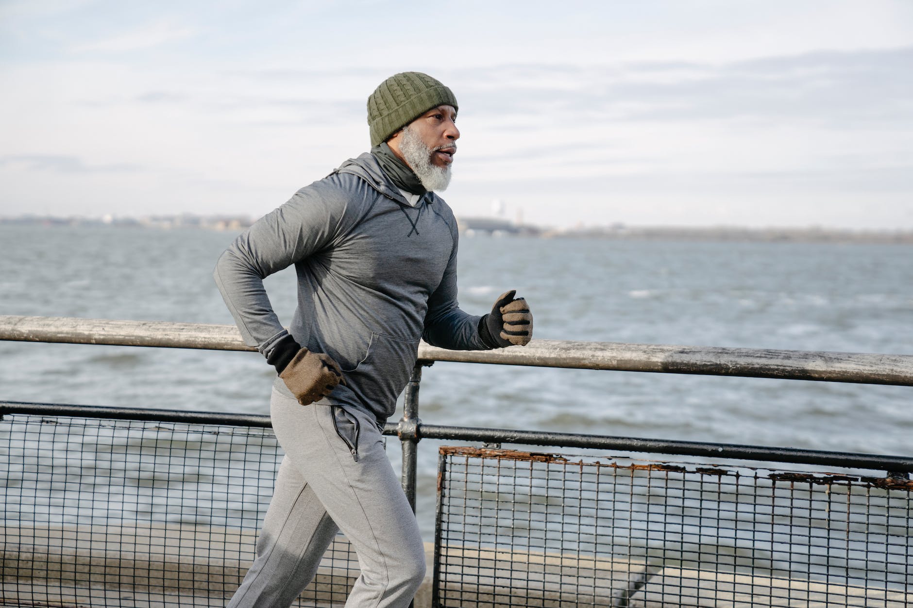 An older man jogging along a path next to a large body of water. He's wearing a grey wool hat, gloves, long-sleeved top and grey pants.