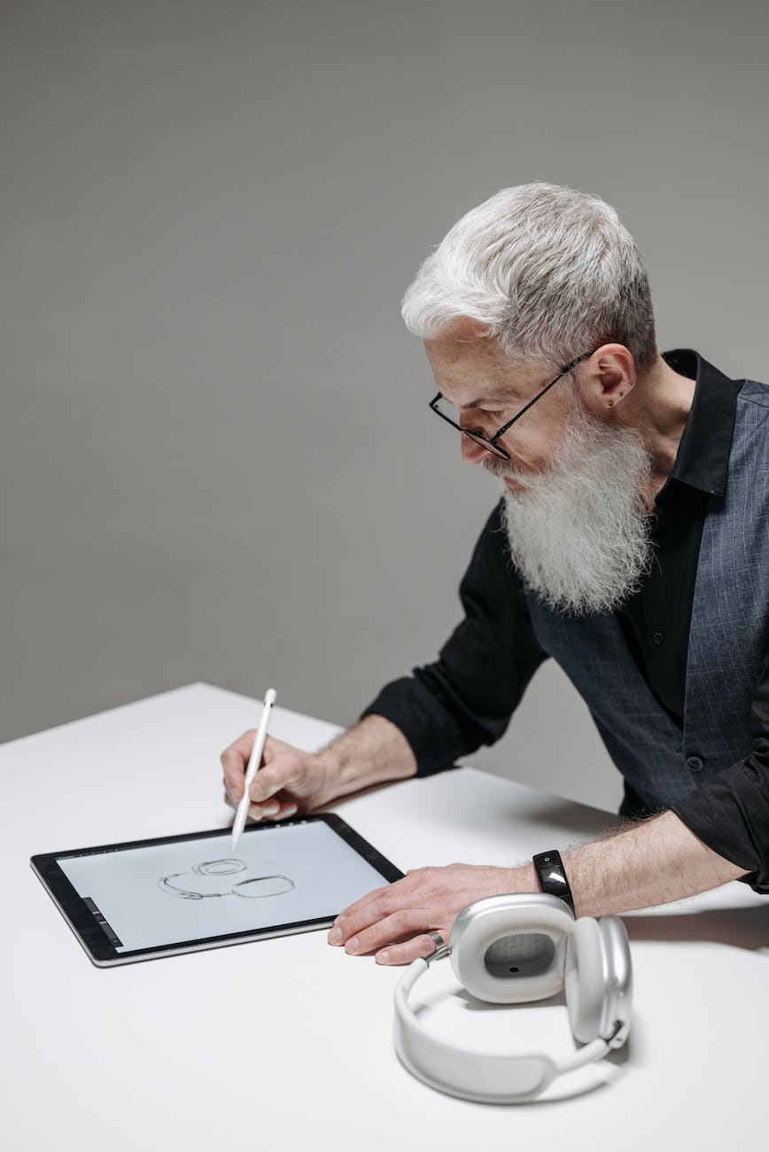 A man with a grey beard and grey hair is standing at a table sketching a pair of white headphones onto a tablet.