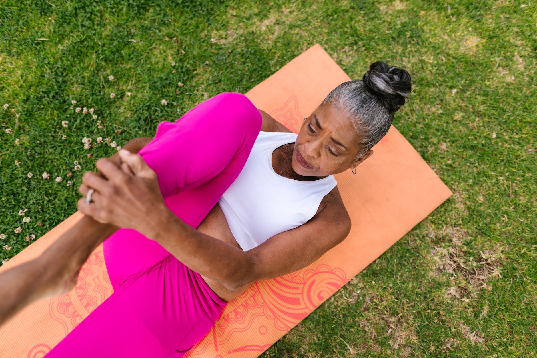 An overhead picture of an older woman exercising on an orange yoga mat. She's wearing pink pants and a white top and holding her right knee to her chest with her hands.