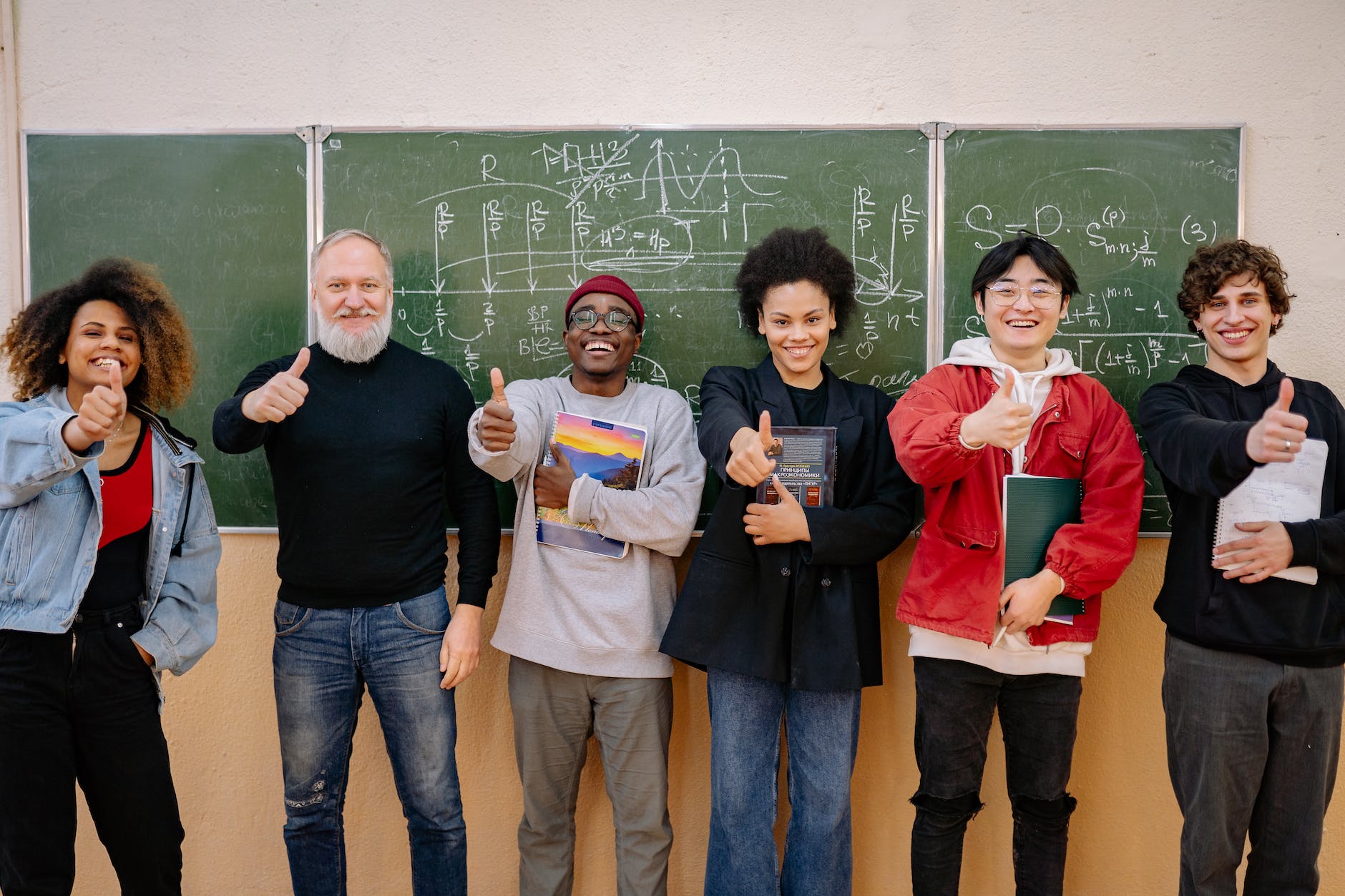Youthful students and a teacher standing in front of a blackboard. They are smiling and giving a thumbs up sign.