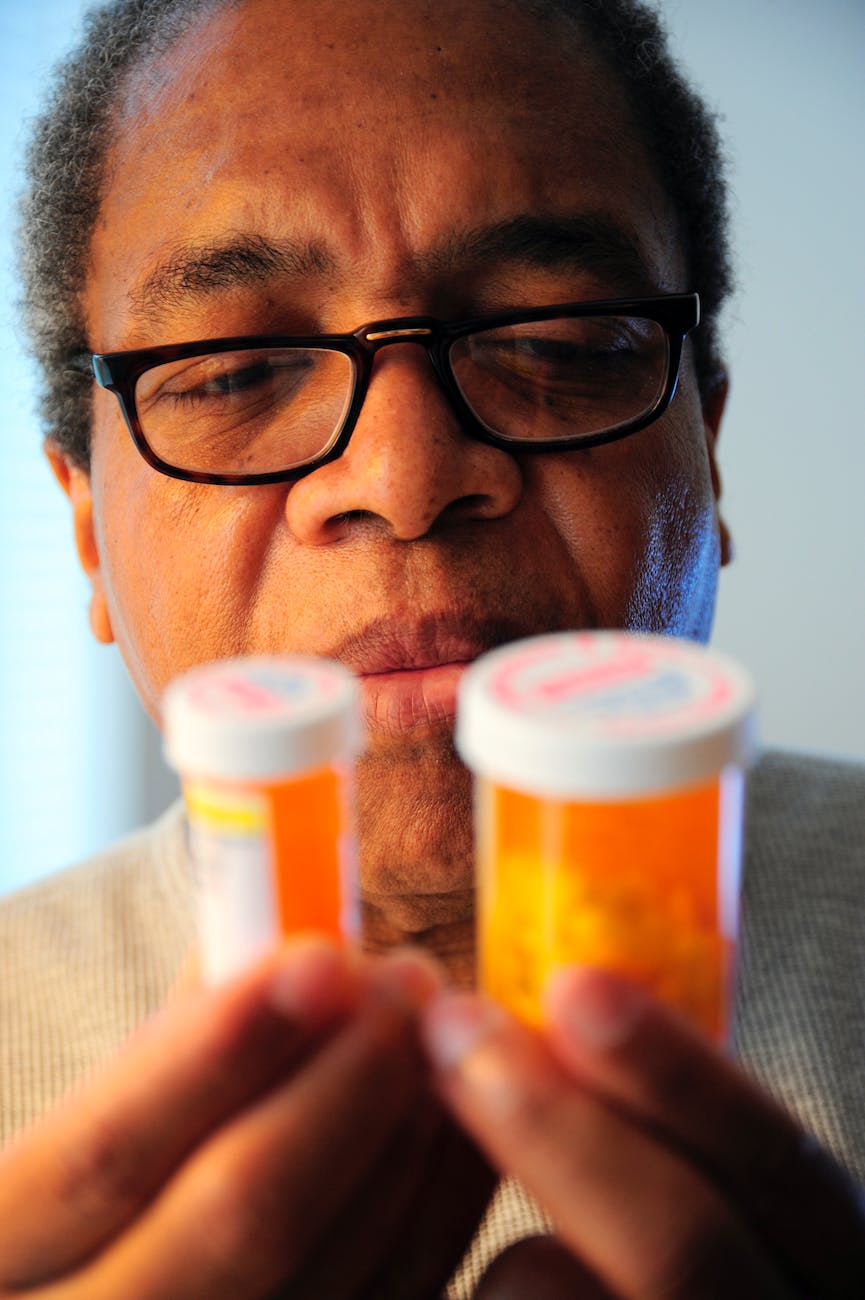 An older man wearing glasses is looking closely at the two medication bottles he is holding.