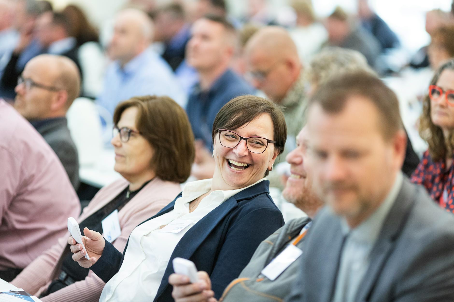 Business people in an audience holding response clickers while sharing a laugh with a a colleague.