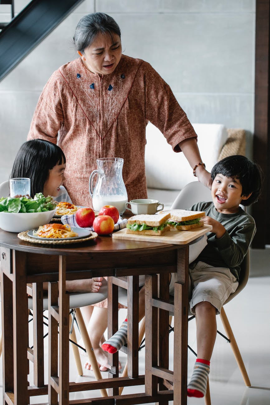 A grandmother stands beside a table where two children are sitting. The table is has milk, apples, sandwiches, salad and spaghetti on it.