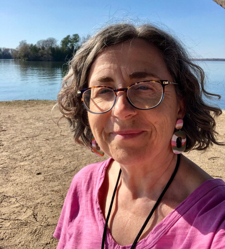 The author smiling standing on a beach on a sunny spring morning with the calm lake behind her.