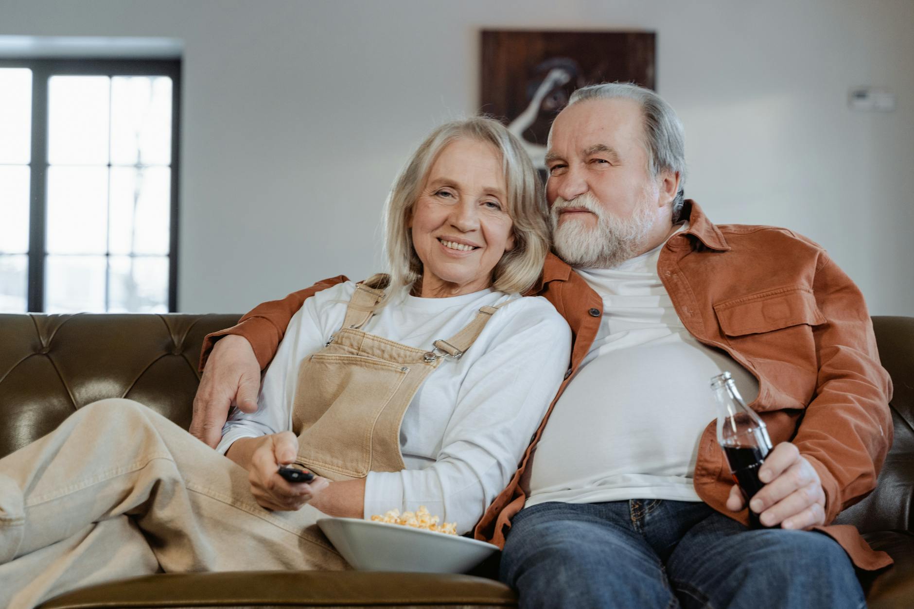 Older couple snuggled on the couch with snacks. The woman is holding the remote and the man holds a drink.
