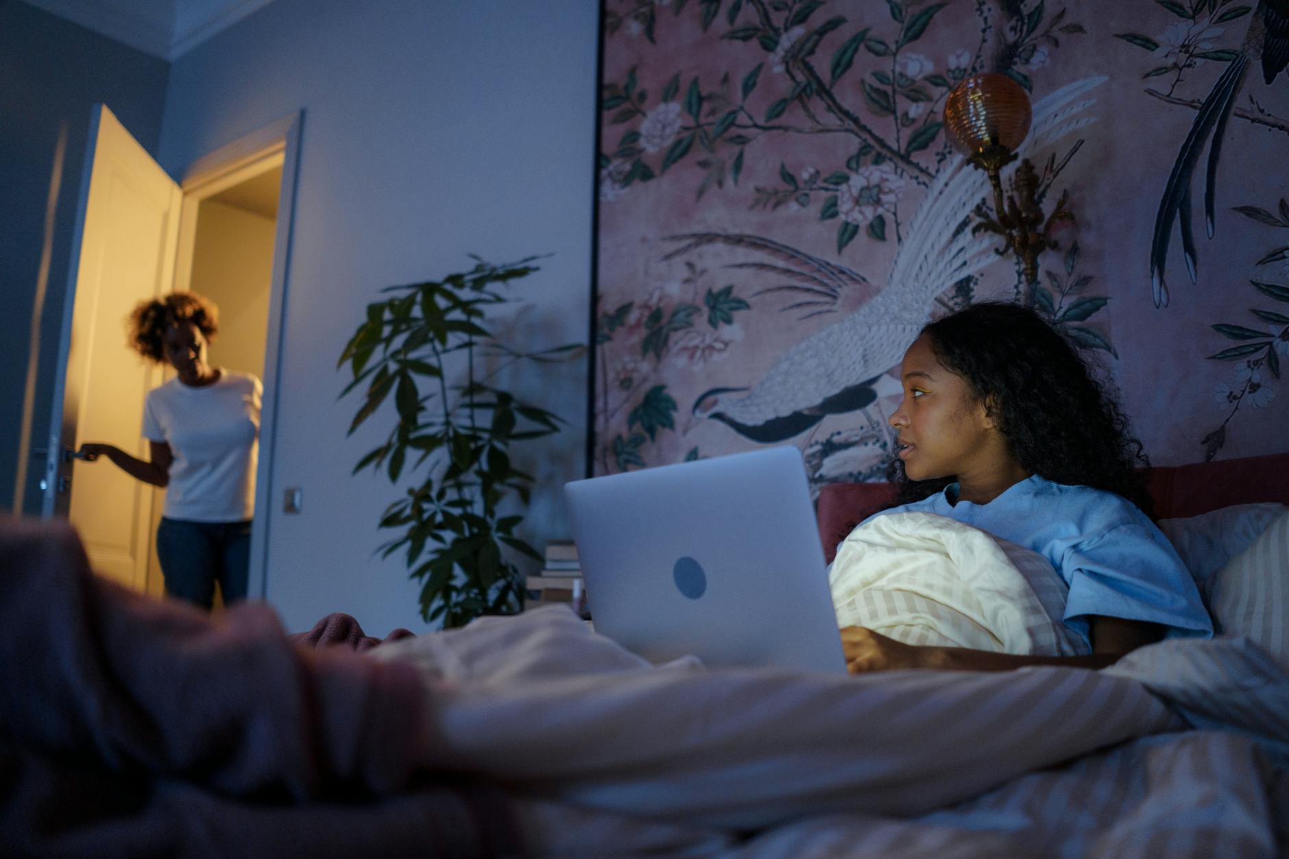A girl is in bed working on her computer at night. A woman has opened the door and is talking with her.
