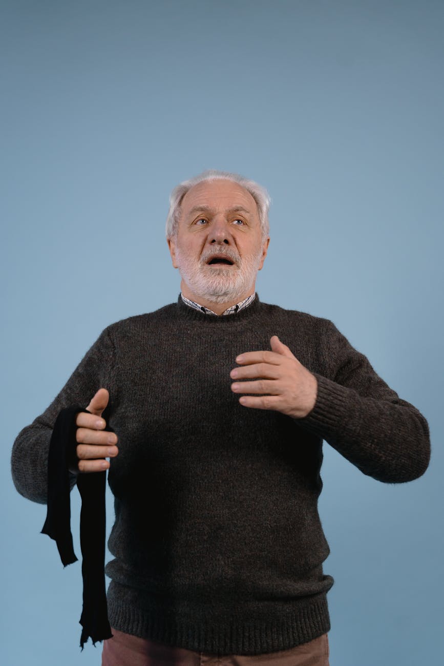 An older man with grey hair and beard is wearing a dark sweater and taking a deep breath.