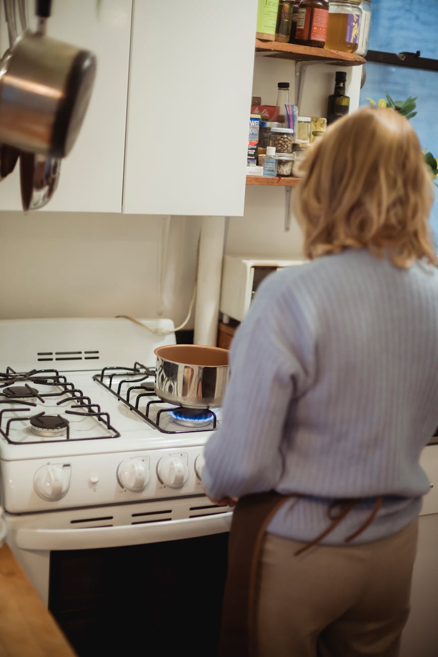 A woman in the kitchen, at the stove, putting a pot on the burner. She has her back to the camera.