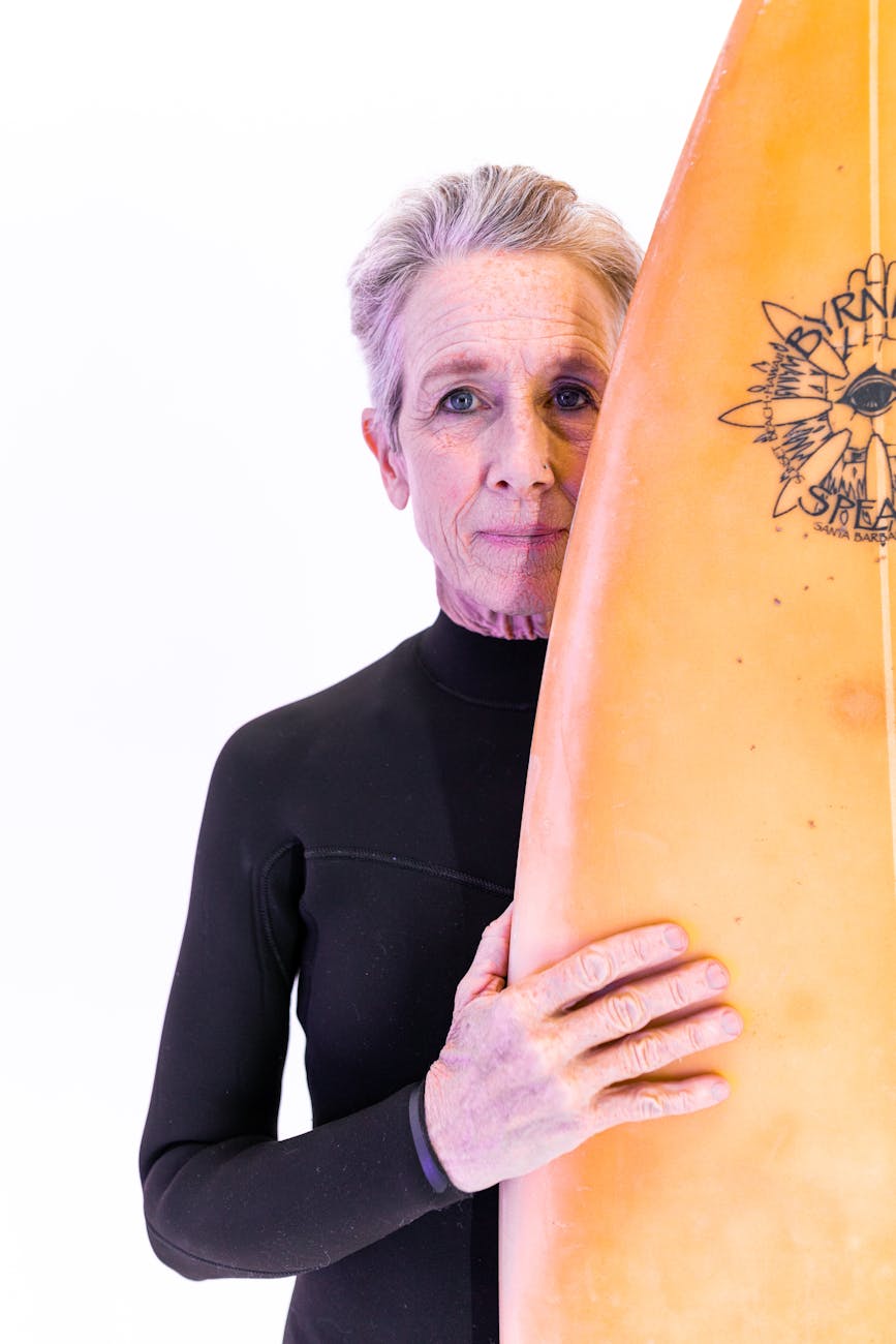 An older person with short grey hair wearing a black wetsuit stands behind a surfboard.