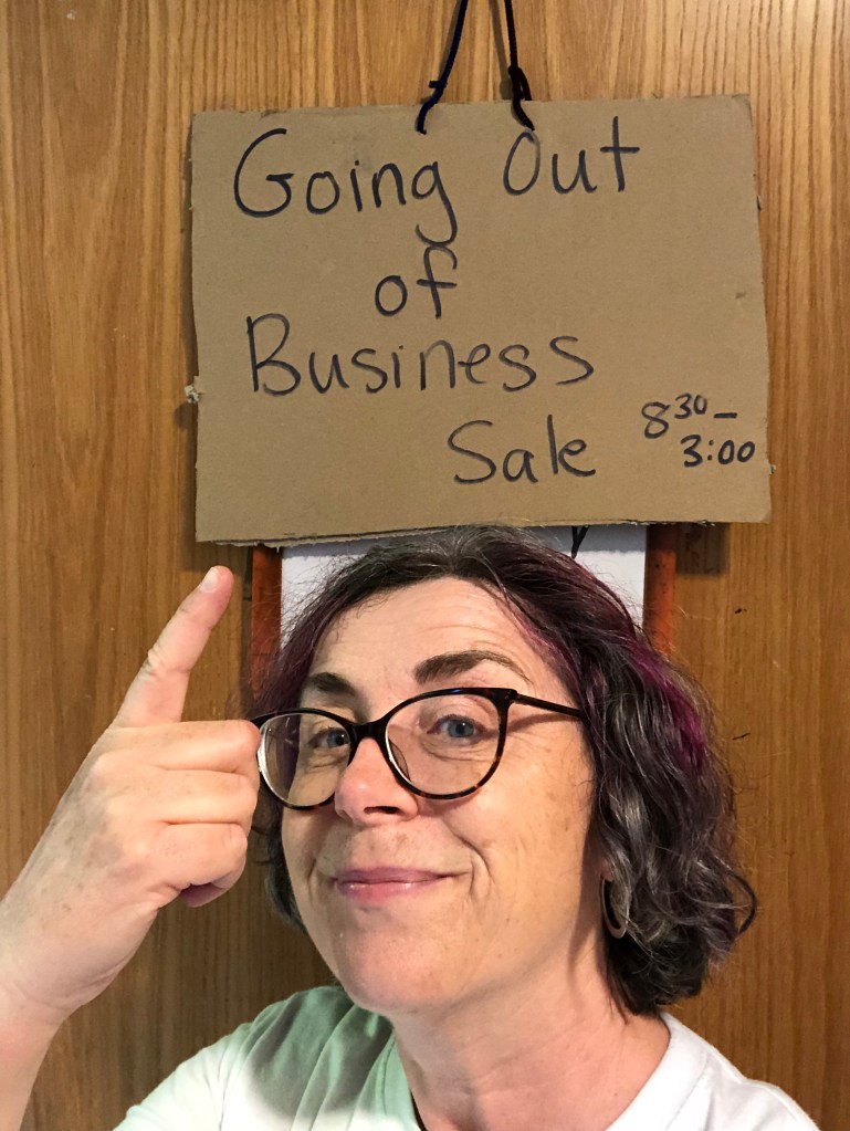 The author smiling and standing in front of her classroom door pointing to a cardboard sign that says, "Going Out Of Business Sale."