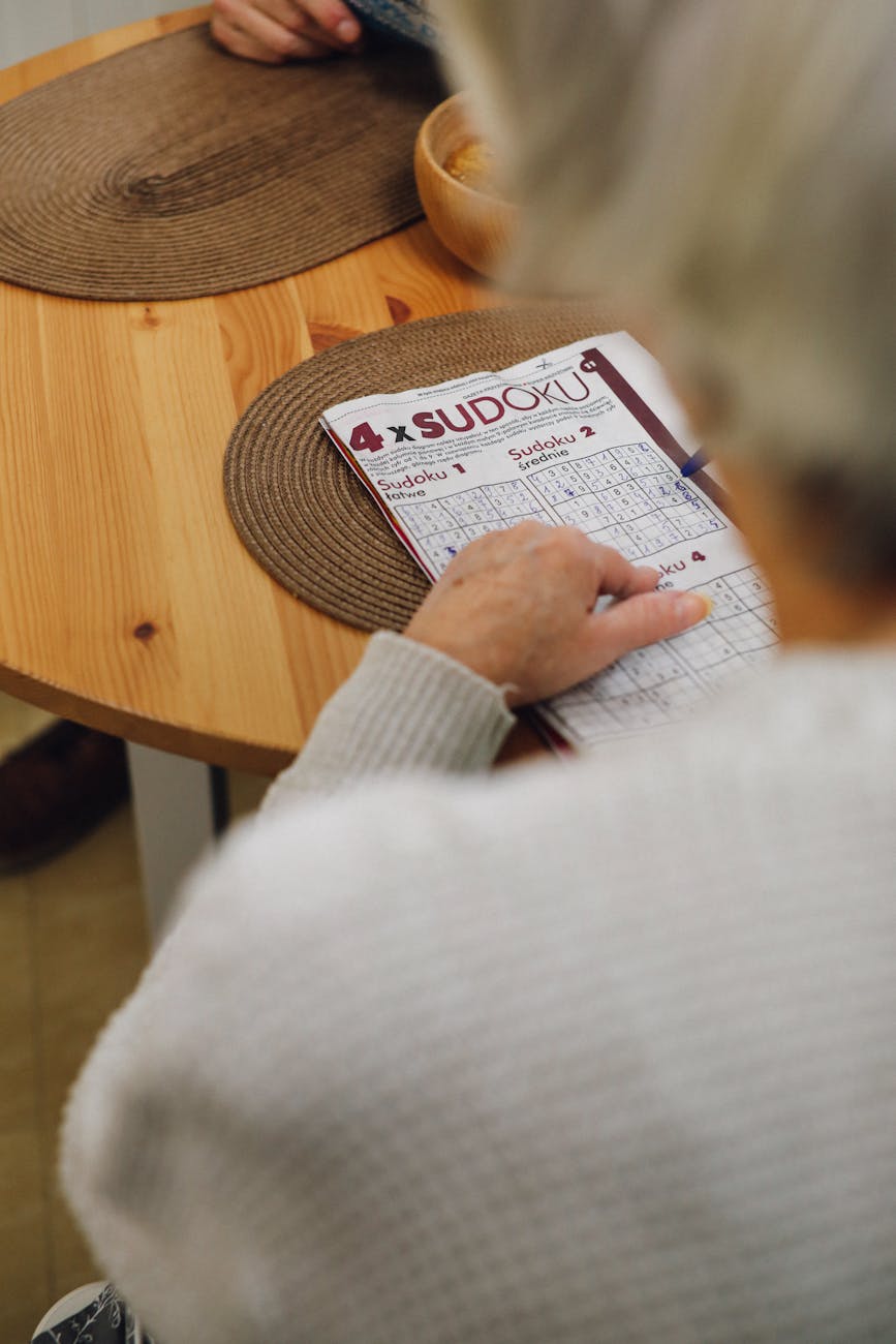 A view over the shoulder of an older person, sitting at a table as they do the sudoku puzzle.