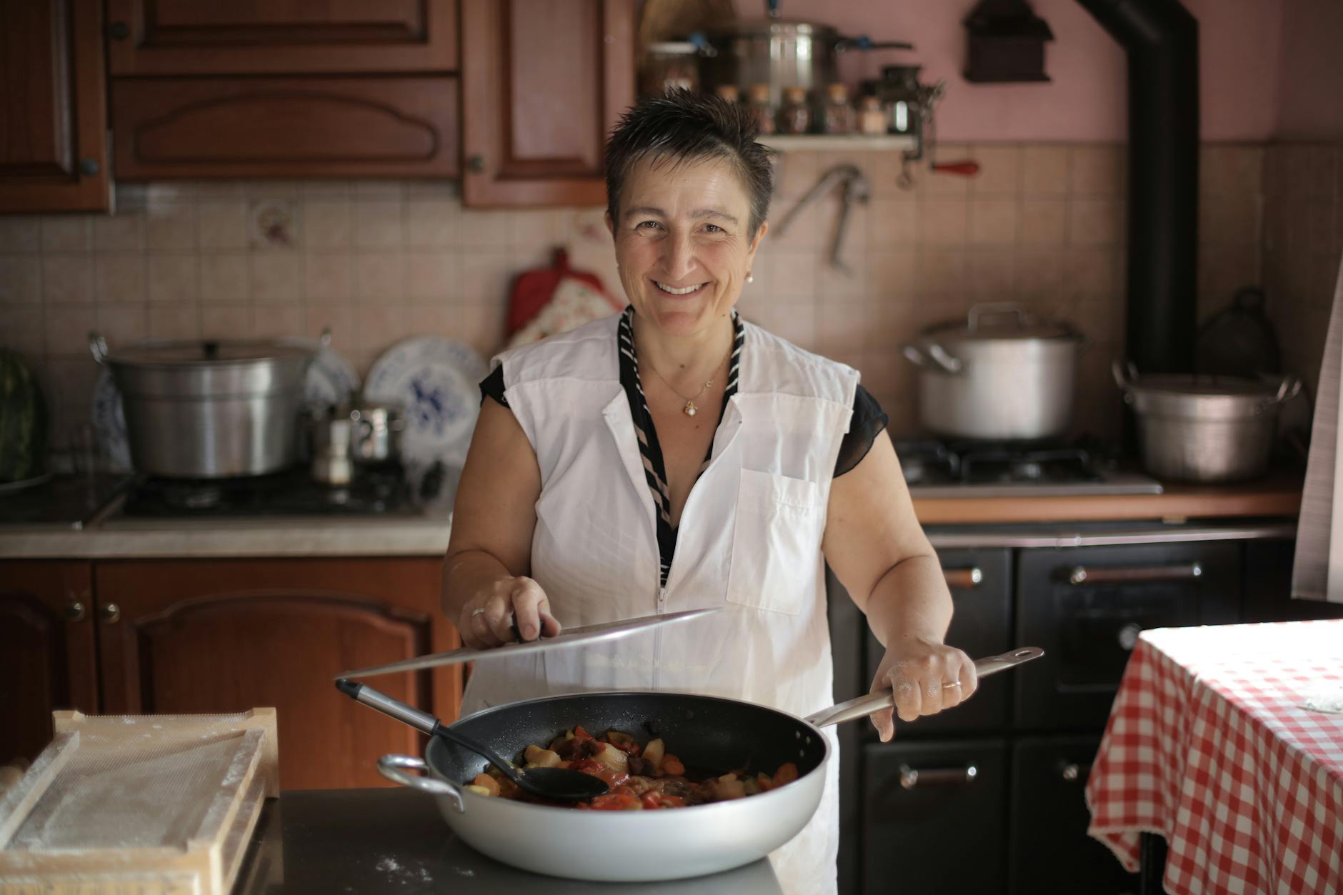 A woman in a kitchen opening the lid of a big skillet containing cooked food. She is smiling.