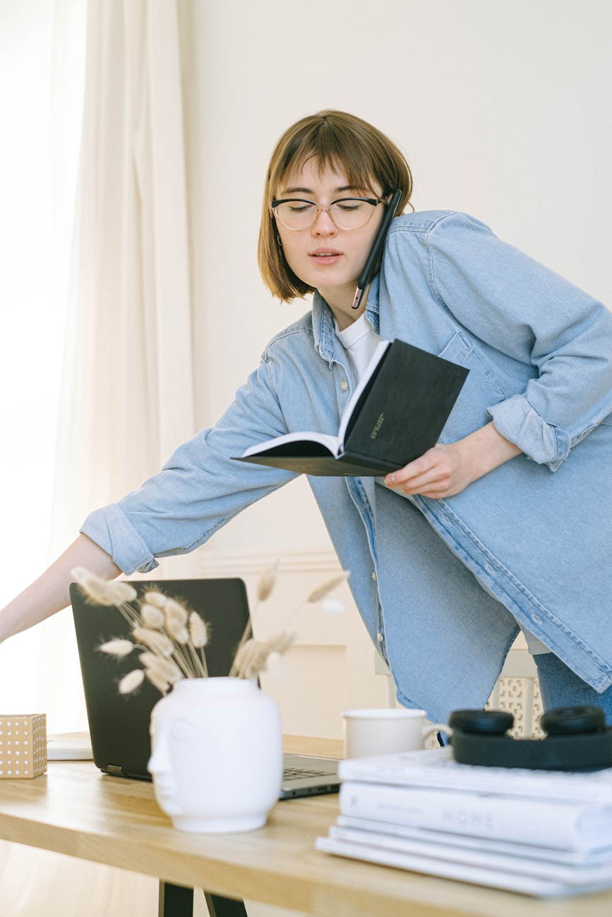 A person reaching for a pen while on the phone and holding a day planner as they stand behind a desk.