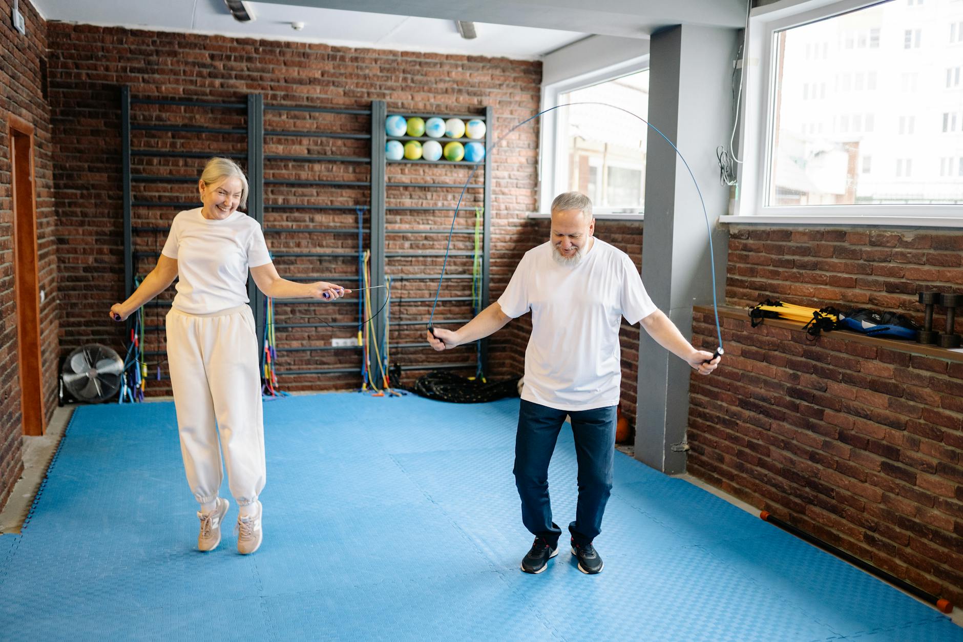 A smiling older woman and a man are skipping rope in a gym with a blue floor.
