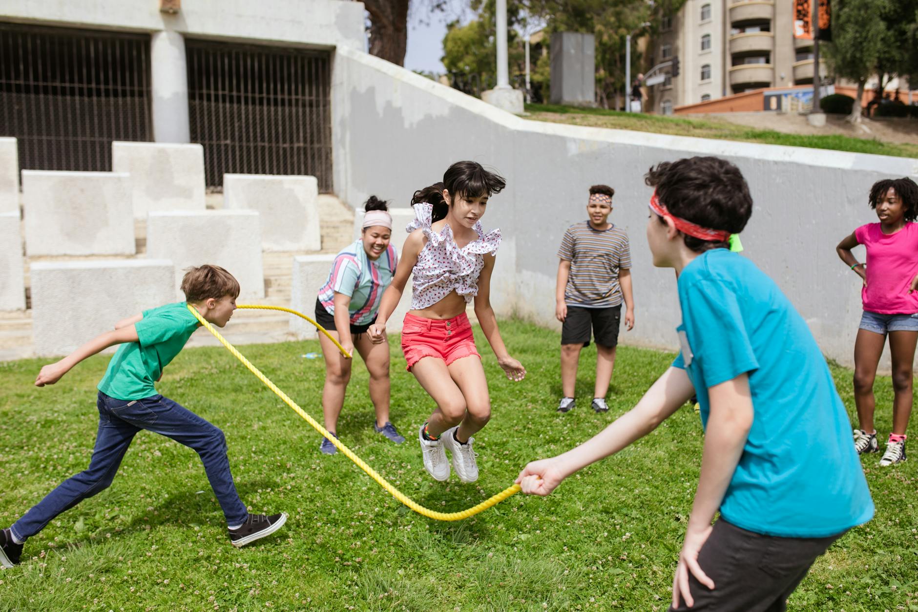Children are holding each end of a long rope and jumping in to skip. They are outside on the grass.