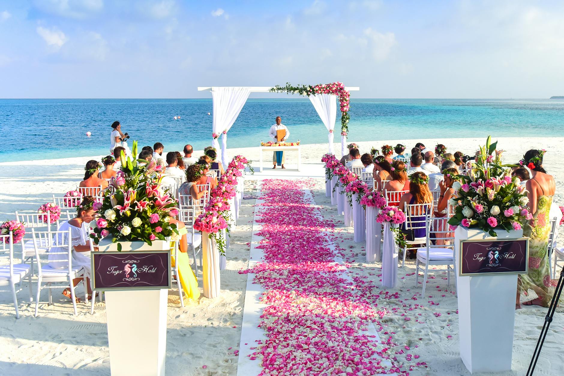 The view down the flower strewn aisle of a beach wedding. The guests are seated facing the water and the officiant is standing in centre.