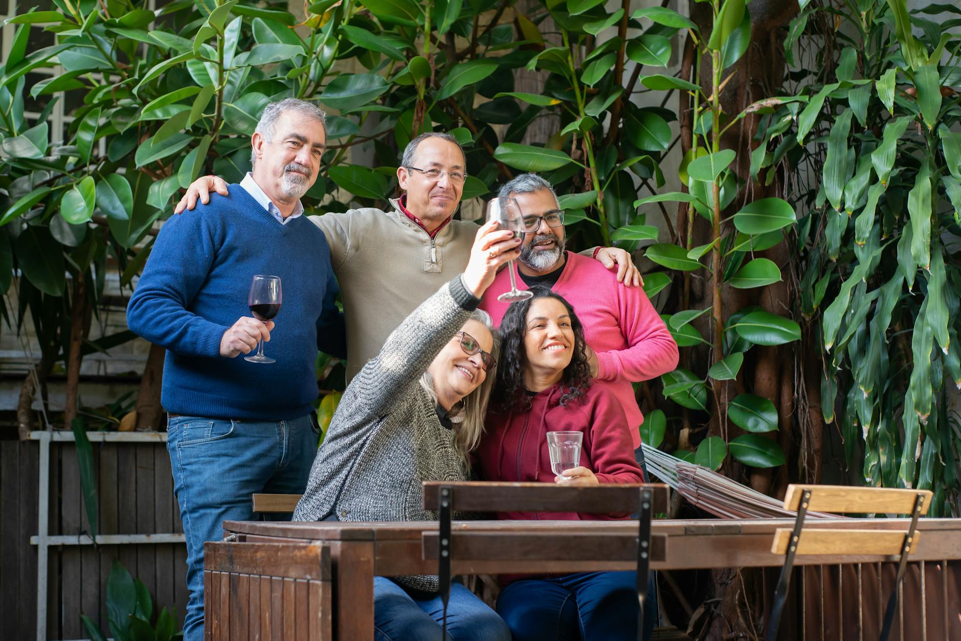 Three men stand behind and two women are seated. Most are holding glasses of wine and smiling at a camera and indicating, "Cheers."