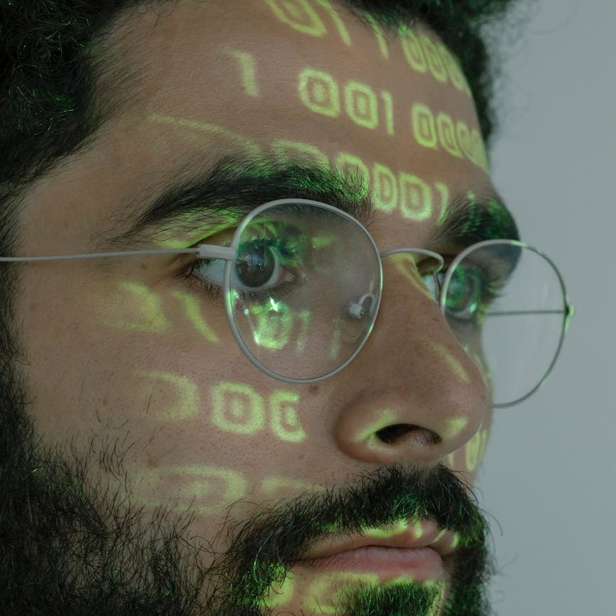 A man's face has computer code reflected on it from the screen he is staring at.