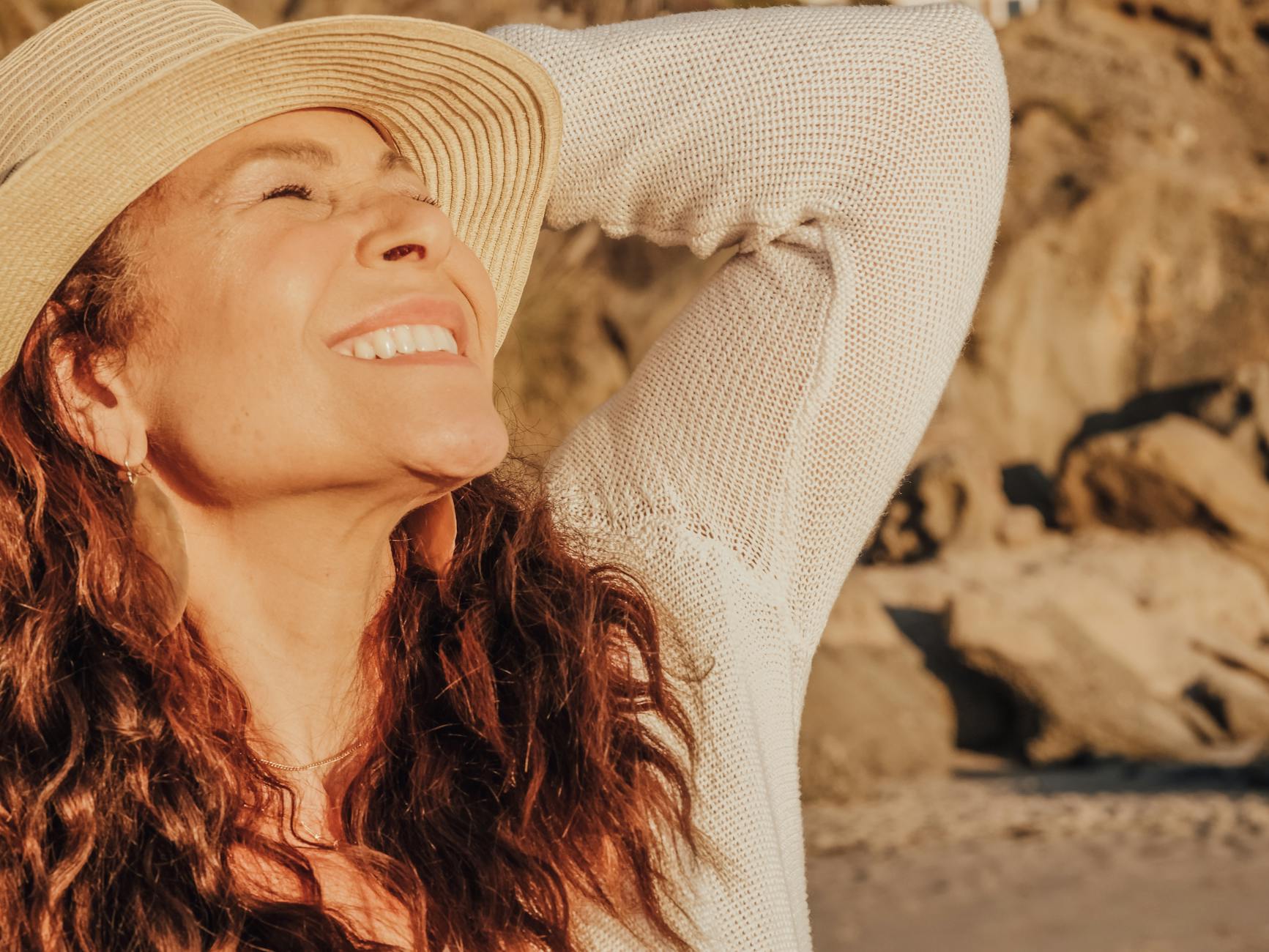 A woman with long dark hair, wearing a straw hat and neutral long-sleaved top, scrunches up her face as she raises it up to the sun. There are mountainous rocks behind her.