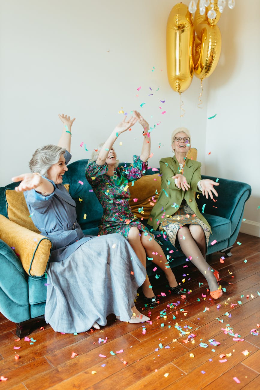 Three midlife women sit on a couch and throw their arms in the air in celebration as confetti falls down on them.