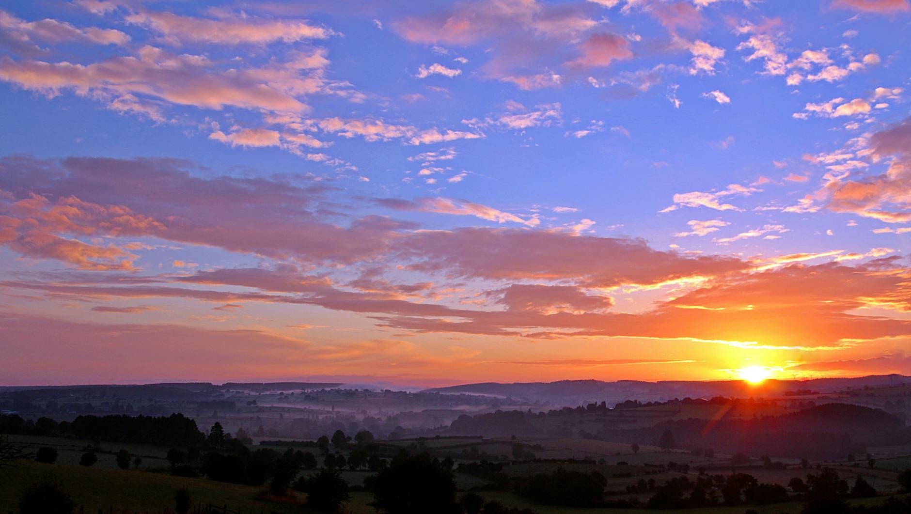 The sun is rising in the distance over the hills and trees. The sky is yellow and blue and the clouds are pink.