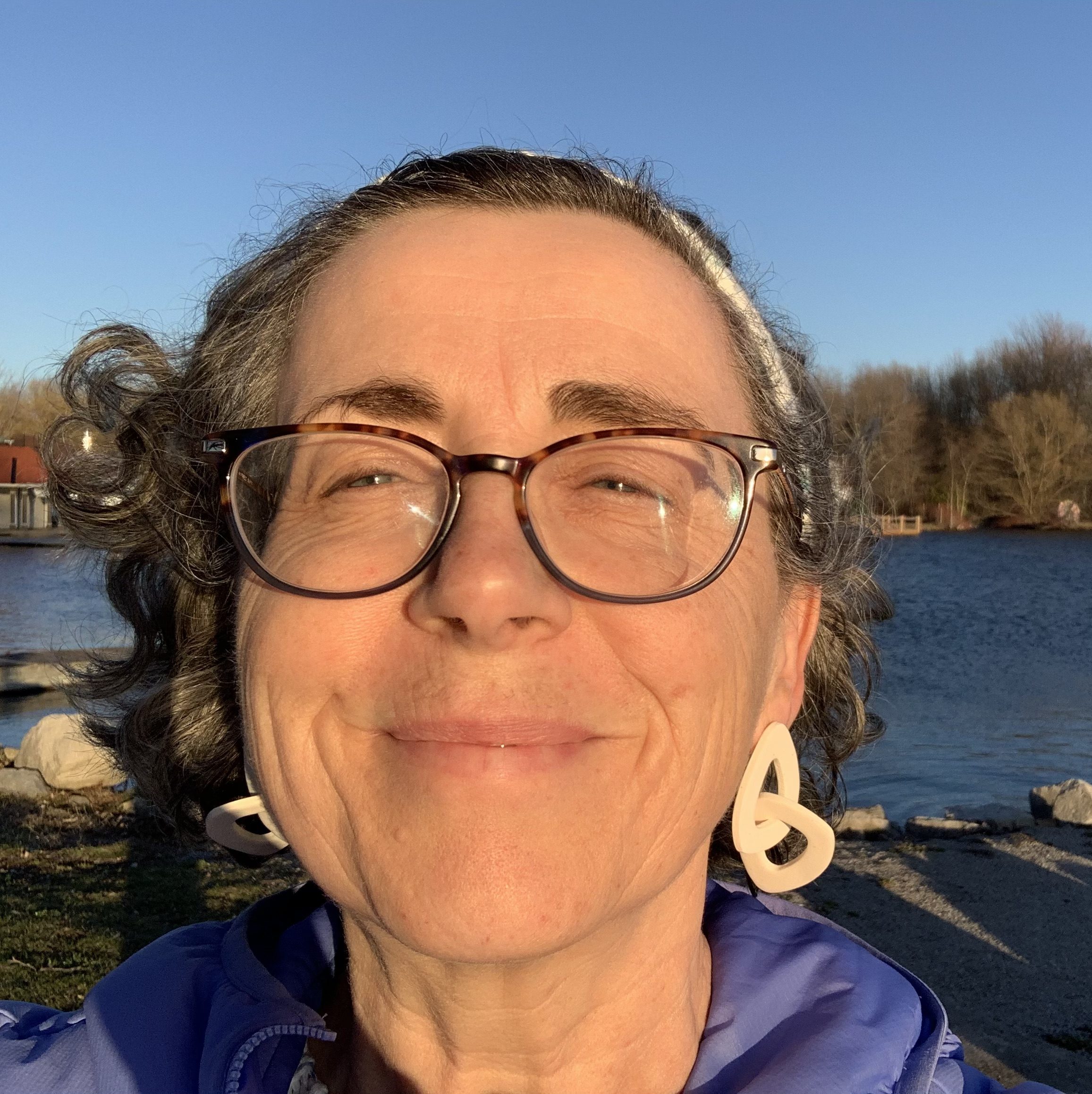 The author smiling with the sun in her face and the lake behind her. She is wearing brown glasses, large white earrings and a blue coat.