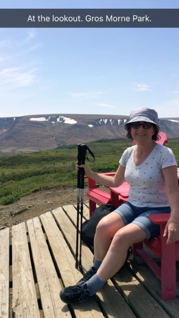 The author sitting on one of two red muskoka chairs and smiling. She is wearing a sunhat, t-shirt and shorts and she is holding her hiking poles in one hand. The text says, "At the lookout. Gros Morne Park."