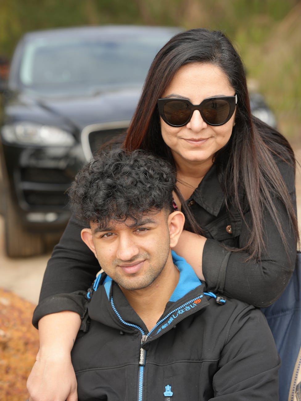 A mom and teen son pose in front of a parked car. The mom is wearing sunglasses and smiling. She has her arm casually resting on her son's shoulder.