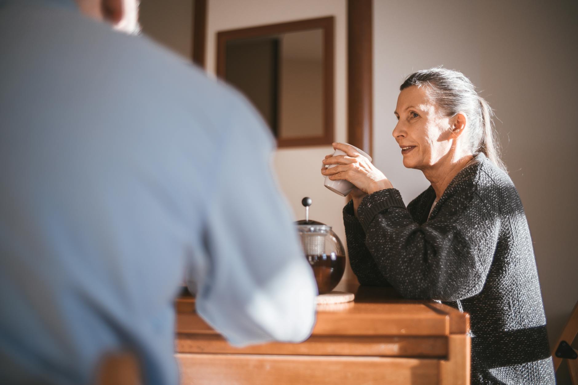 Two people sit at a table in a home drinking coffee. The woman is in profile responding to the other person who has their back to the camera.