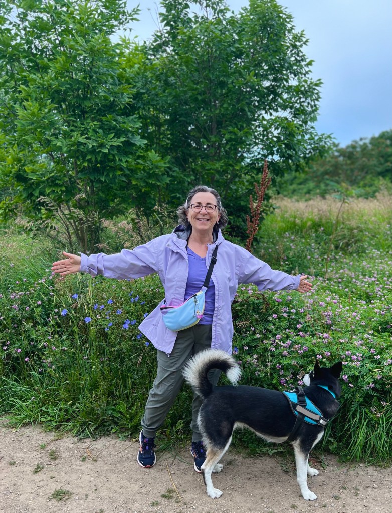 The author standing with arms outstretched in front of a field of spring flowers. There is a husky next to her.