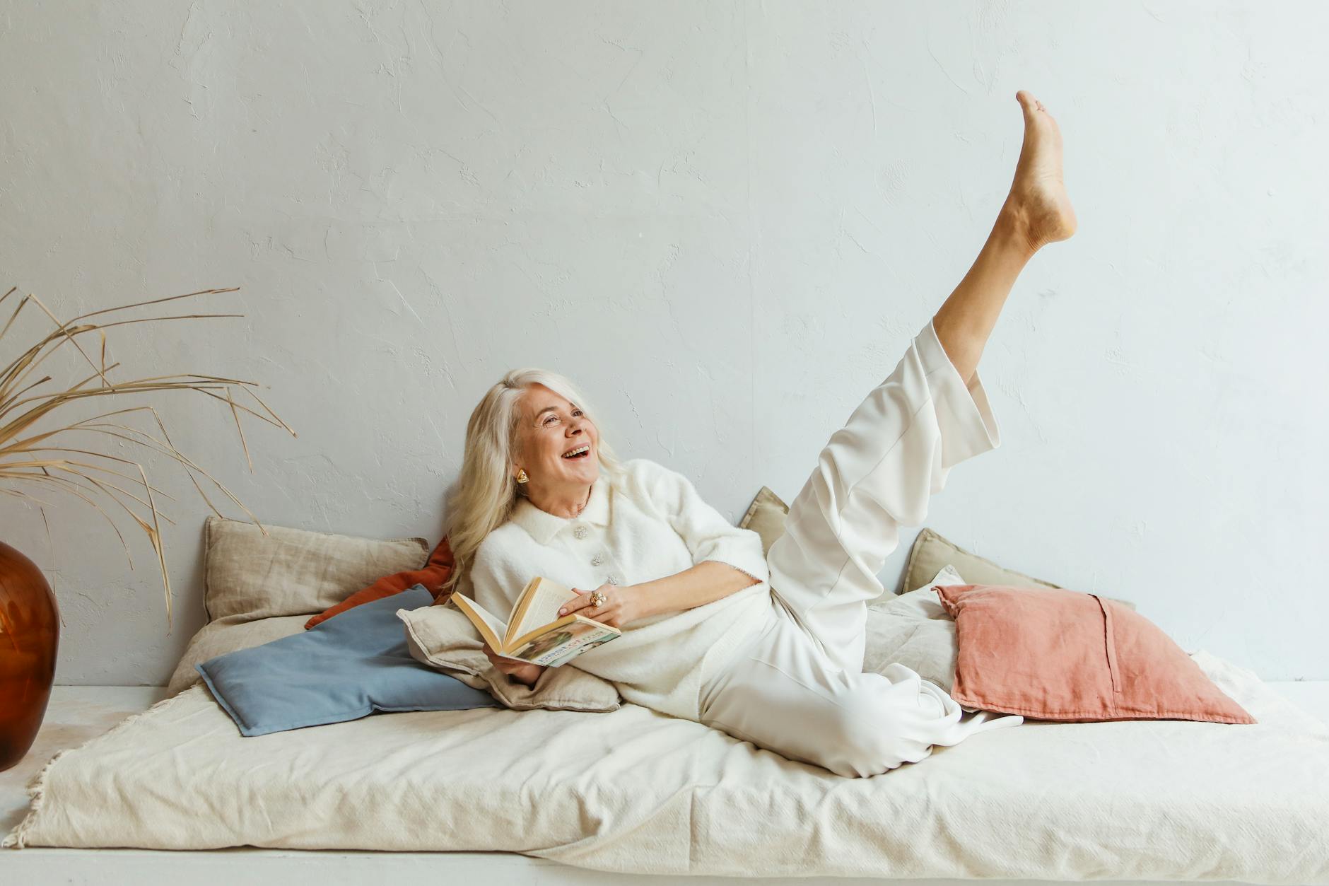 A joyous woman reclining on some pillows pausing while reading to laugh and throw her leg up into the air.