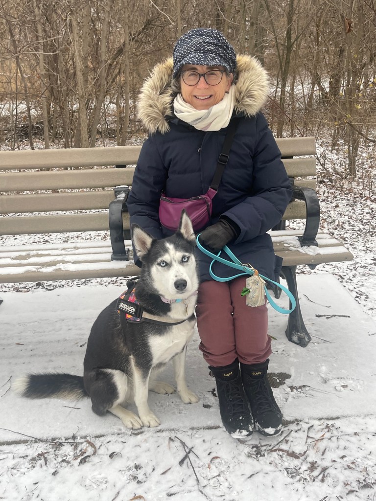 The author sits on a park bench in with a husky at her feet. It's winter and she's wearing a blue parker, blue hat and black boots.