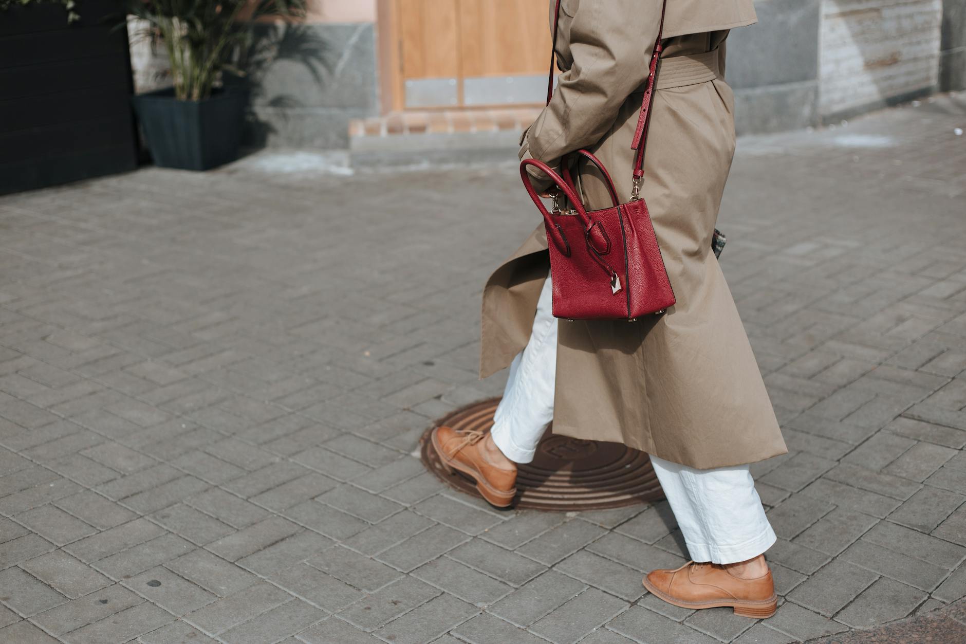 A person in a beige trench coat, wearing white pants, brown flat shoes and a red purse walks purposefully along a city street.
