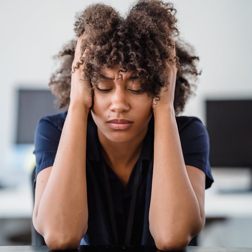 A young person in a navy t-shirt sits with their hands in their hair and their eyes looking downward.