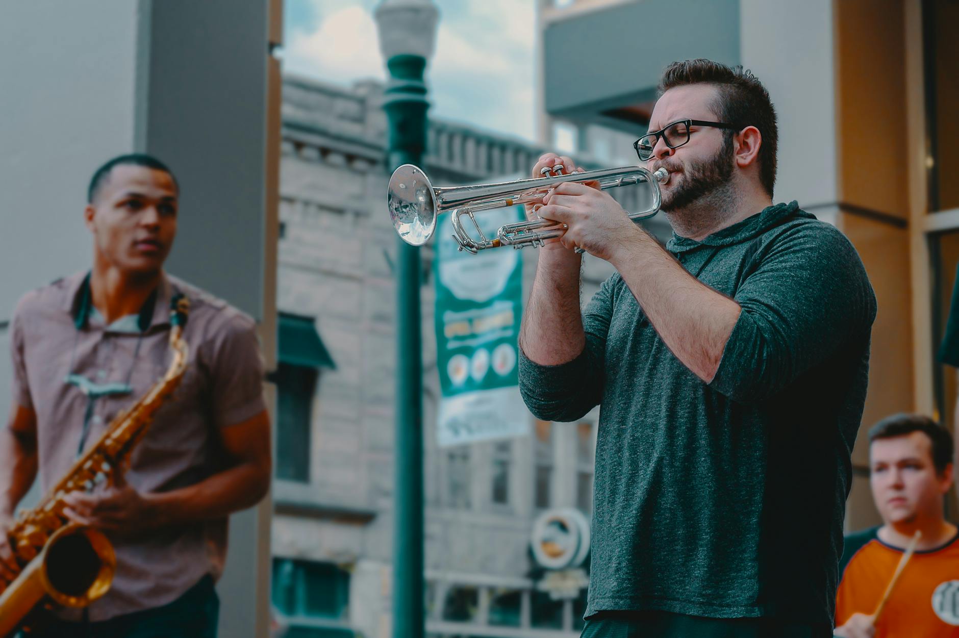 Three men are playing musical instruments. On the left is a saxophone player, in the middle in a green shirt is a trumpet player and in the right at the back is a drummer.
