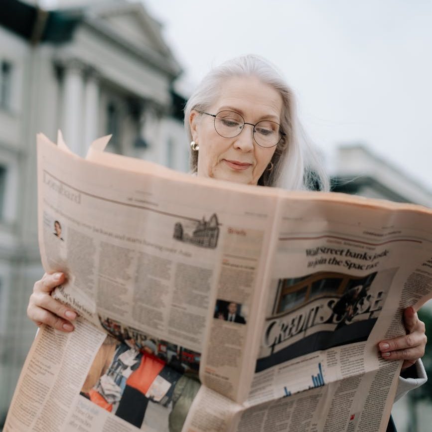 An older woman wearing glasses is reading a newspaper on the steps of a civil building.