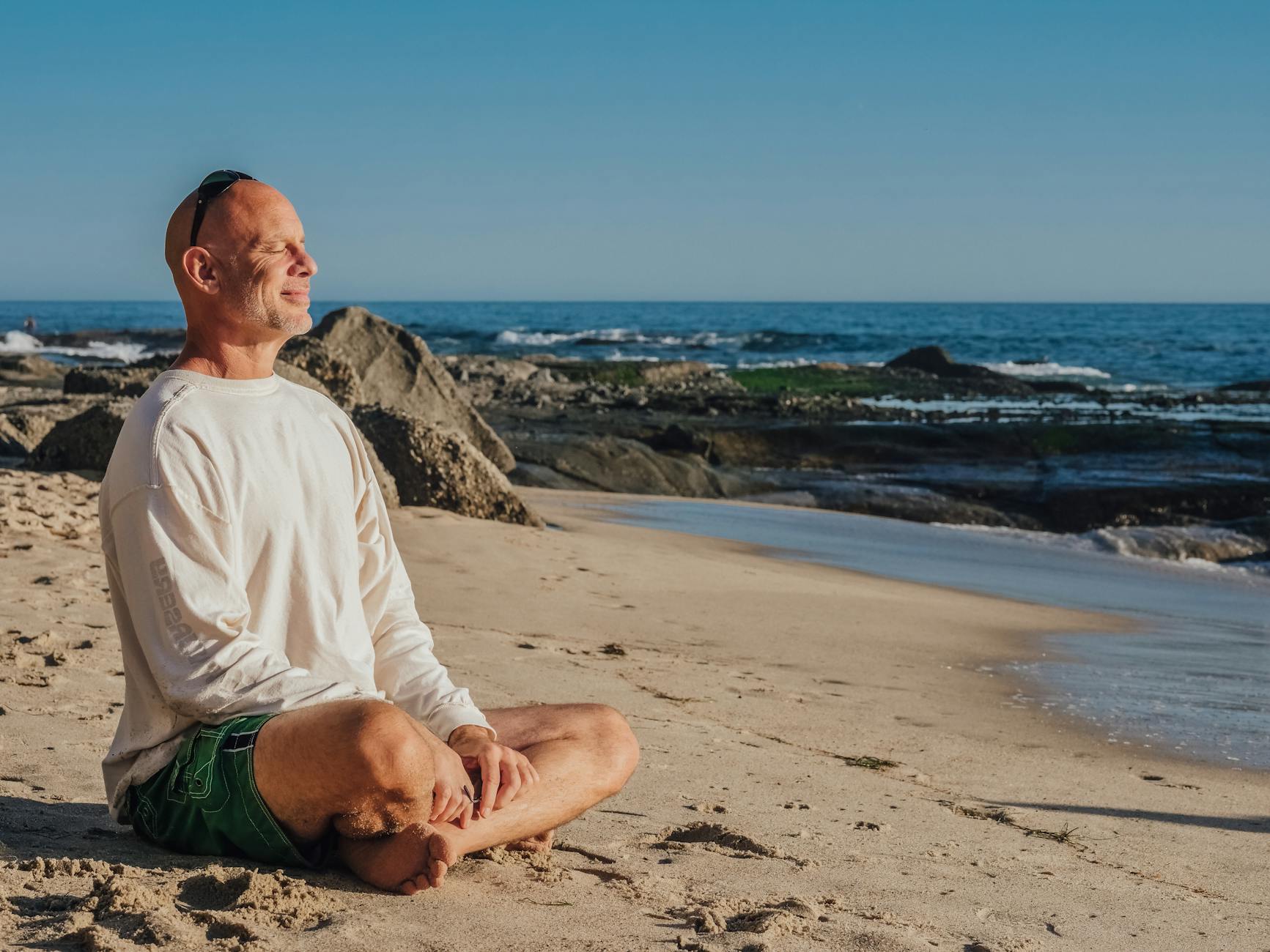 An older man is smiling contently with his eyes closed while sitting cross legged on the beach facing the water.